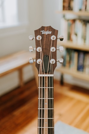 A close-up view of a guitar headstock, featuring tuning pegs and strings. The guitar is placed indoors with a blurred background showing wooden floors and a bookshelf.