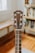 A close-up view of a guitar headstock, featuring tuning pegs and strings. The guitar is placed indoors with a blurred background showing wooden floors and a bookshelf.
