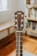 A close-up view of a guitar headstock, featuring tuning pegs and strings. The guitar is placed indoors with a blurred background showing wooden floors and a bookshelf.