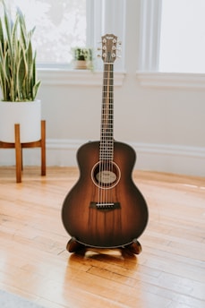 A brown acoustic guitar stands upright on a wooden floor in a well-lit room. The guitar is framed by large windows and a potted plant placed in the background, creating a serene and aesthetically pleasing atmosphere.