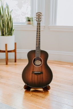 A brown acoustic guitar stands upright on a wooden floor in a well-lit room. The guitar is framed by large windows and a potted plant placed in the background, creating a serene and aesthetically pleasing atmosphere.