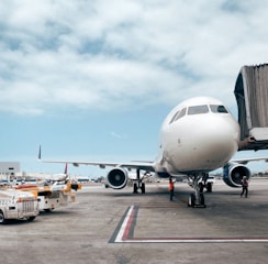 A large commercial airplane is stationed on the tarmac at an airport. The aircraft is connected to a jet bridge, indicating that it is either boarding or disembarking passengers. Ground vehicles, including tugs and baggage carts, are visible nearby, along with airport personnel in reflective vests attending to tasks around the plane. The sky is partially cloudy, adding to the busy atmosphere of the airport setting.