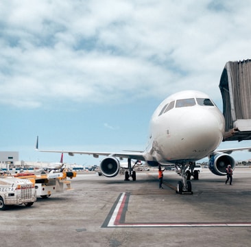 A large commercial airplane is stationed on the tarmac at an airport. The aircraft is connected to a jet bridge, indicating that it is either boarding or disembarking passengers. Ground vehicles, including tugs and baggage carts, are visible nearby, along with airport personnel in reflective vests attending to tasks around the plane. The sky is partially cloudy, adding to the busy atmosphere of the airport setting.