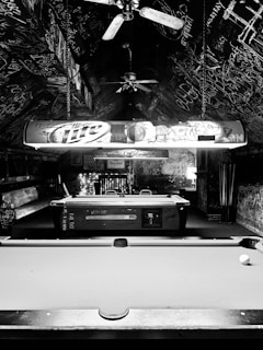A black and white photo of a room with a pool table in the foreground. The ceiling and walls are covered in graffiti and writing, adding an urban and edgy atmosphere. Ceiling fans are present, and a light fixture with branding hangs over the pool table. The room has a somewhat cluttered and chaotic feel, with additional seating or furniture visible in the background.