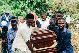 Several people are carrying a wooden coffin with brass handles in an outdoor setting surrounded by greenery. The group appears somber and focused on the task at hand.