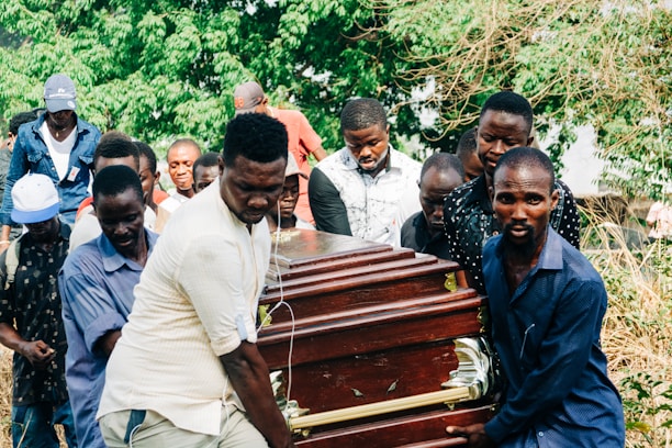 Several people are carrying a wooden coffin with brass handles in an outdoor setting surrounded by greenery. The group appears somber and focused on the task at hand.