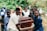 Several people are carrying a wooden coffin with brass handles in an outdoor setting surrounded by greenery. The group appears somber and focused on the task at hand.