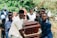Several people are carrying a wooden coffin with brass handles in an outdoor setting surrounded by greenery. The group appears somber and focused on the task at hand.
