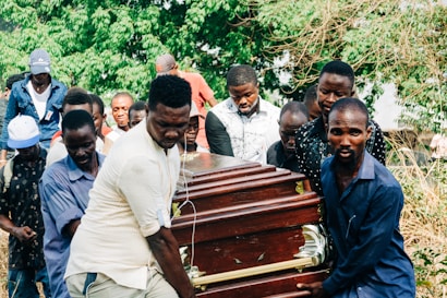 Several people are carrying a wooden coffin with brass handles in an outdoor setting surrounded by greenery. The group appears somber and focused on the task at hand.