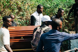 A group of men are carrying a wooden coffin through a lush, green area. The men are dressed casually, and one wears a white cap while another has a backpack. The background features dense greenery, suggesting a forested or rural setting.