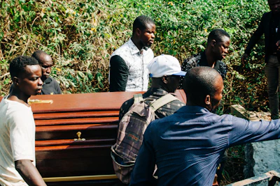 A group of men are carrying a wooden coffin through a lush, green area. The men are dressed casually, and one wears a white cap while another has a backpack. The background features dense greenery, suggesting a forested or rural setting.