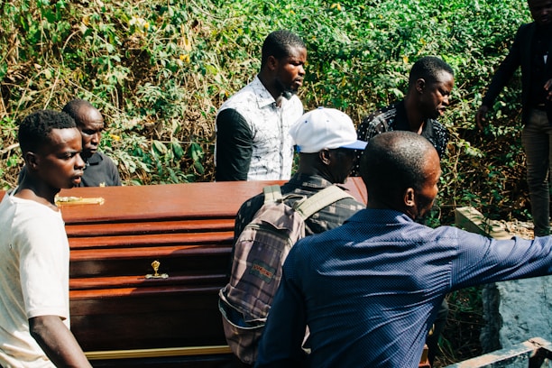 A group of men are carrying a wooden coffin through a lush, green area. The men are dressed casually, and one wears a white cap while another has a backpack. The background features dense greenery, suggesting a forested or rural setting.