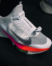 A close-up view of a modern athletic shoe featuring a sleek white and gray design with a prominent locking mechanism labeled 'LOCK' and text 'FLYEASE'. The shoe has colorful accents including a bright pink and orange sole.