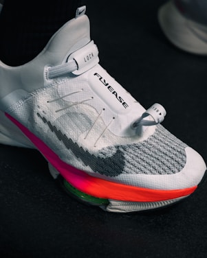 A close-up view of a modern athletic shoe featuring a sleek white and gray design with a prominent locking mechanism labeled 'LOCK' and text 'FLYEASE'. The shoe has colorful accents including a bright pink and orange sole.