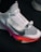A close-up view of a modern athletic shoe featuring a sleek white and gray design with a prominent locking mechanism labeled 'LOCK' and text 'FLYEASE'. The shoe has colorful accents including a bright pink and orange sole.