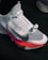 A close-up view of a modern athletic shoe featuring a sleek white and gray design with a prominent locking mechanism labeled 'LOCK' and text 'FLYEASE'. The shoe has colorful accents including a bright pink and orange sole.