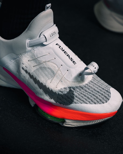 A close-up view of a modern athletic shoe featuring a sleek white and gray design with a prominent locking mechanism labeled 'LOCK' and text 'FLYEASE'. The shoe has colorful accents including a bright pink and orange sole.