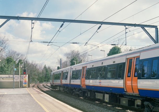 A train is passing through a station on a clear day. The platform is empty, with signs and a red signal visible. Overhead wires and track curves add to the scene's detail, surrounded by trees and a partly cloudy sky.