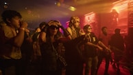A group of people enjoying a lively event in a dimly lit room with vibrant lighting. Some are dancing, while others are wearing sunglasses and expressive clothing, suggesting a nightclub or concert setting.