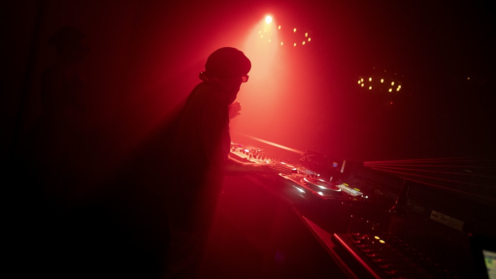 A silhouetted DJ stands behind a turntable setup in a dimly lit room, illuminated by red light and surrounded by smoke or fog. The atmosphere is moody and immersive, heightened by shadows and reflections on the equipment.