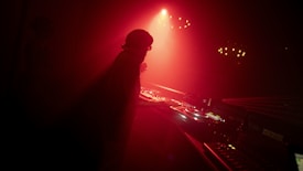 A silhouetted DJ stands behind a turntable setup in a dimly lit room, illuminated by red light and surrounded by smoke or fog. The atmosphere is moody and immersive, heightened by shadows and reflections on the equipment.