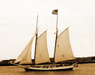 A large, classic sailing ship with three sails unfurled is moving across a body of water. The ship has an American flag hoisted on the mast, and there is a group of people visible on the deck. The background features a coastline with some industrial buildings.