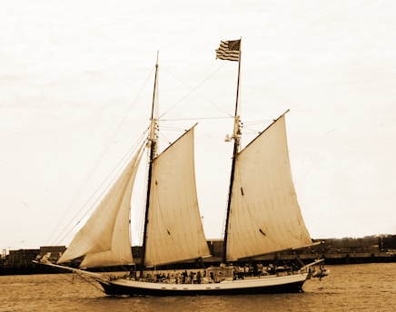 A large, classic sailing ship with three sails unfurled is moving across a body of water. The ship has an American flag hoisted on the mast, and there is a group of people visible on the deck. The background features a coastline with some industrial buildings.