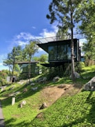 A modern glass house is situated on a lush, green hillside surrounded by trees. The structure features large windows reflecting the sky and the natural scenery. Bright blue sky with scattered clouds can be seen above, creating a serene and peaceful environment. Rocks and well-maintained grass cover the slope leading up to the house.
