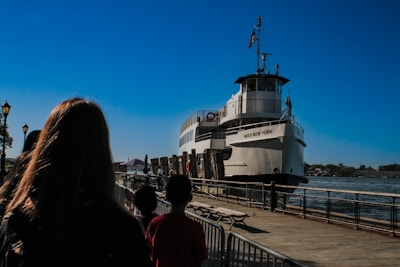 A large white ferry named 'Miss New York' is docked by the pier, with several people standing on the walkway beside it. The sky is clear and blue, and the waterfront is lined with railings and benches. Trees and lamp posts are visible in the background.