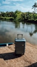 A hard-shell suitcase stands on a stone surface near a tranquil body of water, surrounded by lush green foliage and palm trees under a blue sky.