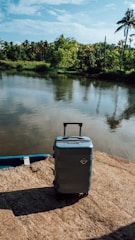 A hard-shell suitcase stands on a stone surface near a tranquil body of water, surrounded by lush green foliage and palm trees under a blue sky.