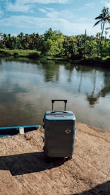 A hard-shell suitcase stands on a stone surface near a tranquil body of water, surrounded by lush green foliage and palm trees under a blue sky.