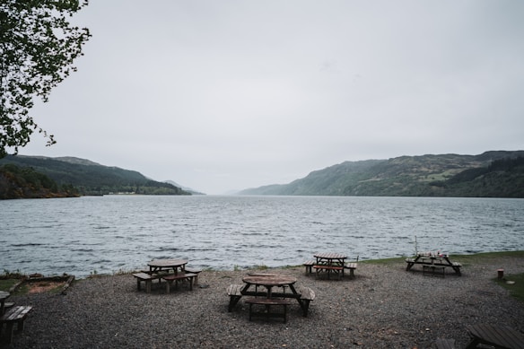 A peaceful lakeside view with several wooden picnic tables scattered on a gravel area by the water's edge. The lake is surrounded by gentle hills covered with lush greenery. The sky is overcast, contributing to a calm and serene atmosphere. A few trees frame the scene, adding a touch of nature to the setting.