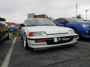 A white Honda car with personalized license plate 'El Casper' is parked in a row with other vehicles. The car features distinct headlights and is located in an outdoor parking lot. Other cars, including a blue vehicle, are adjacent. Background structures and a cloudy sky are visible.