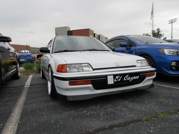 A white Honda car with personalized license plate 'El Casper' is parked in a row with other vehicles. The car features distinct headlights and is located in an outdoor parking lot. Other cars, including a blue vehicle, are adjacent. Background structures and a cloudy sky are visible.