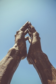 Two large hands made of a dark, textured material are raised against a clear blue sky, with the fingers intertwined in a gesture of prayer or unity.