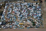 An aerial view of a large junkyard filled with numerous parked vehicles, mainly consisting of trucks, vans, and buses. The vehicles are arranged in a seemingly haphazard manner with dirt paths weaving through them. The area is surrounded by a fence, and there are patches of vegetation visible within and around the yard.