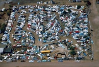 An aerial view of a large junkyard filled with numerous parked vehicles, mainly consisting of trucks, vans, and buses. The vehicles are arranged in a seemingly haphazard manner with dirt paths weaving through them. The area is surrounded by a fence, and there are patches of vegetation visible within and around the yard.