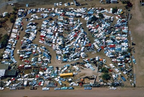 An aerial view of a large junkyard filled with numerous parked vehicles, mainly consisting of trucks, vans, and buses. The vehicles are arranged in a seemingly haphazard manner with dirt paths weaving through them. The area is surrounded by a fence, and there are patches of vegetation visible within and around the yard.
