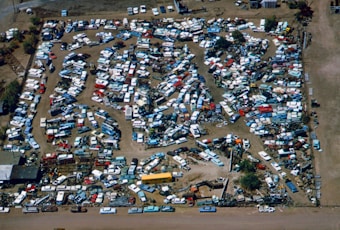 An aerial view of a large junkyard filled with numerous parked vehicles, mainly consisting of trucks, vans, and buses. The vehicles are arranged in a seemingly haphazard manner with dirt paths weaving through them. The area is surrounded by a fence, and there are patches of vegetation visible within and around the yard.