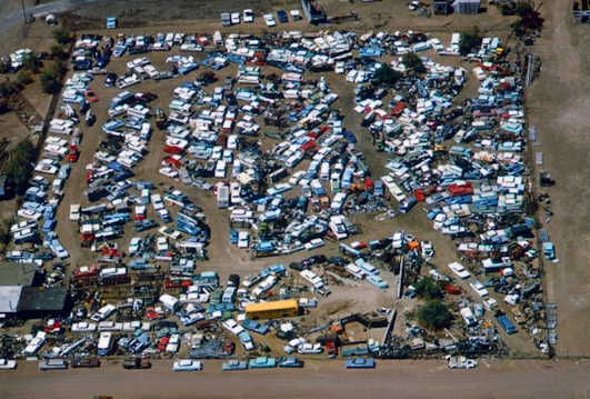 An aerial view of a large junkyard filled with numerous parked vehicles, mainly consisting of trucks, vans, and buses. The vehicles are arranged in a seemingly haphazard manner with dirt paths weaving through them. The area is surrounded by a fence, and there are patches of vegetation visible within and around the yard.