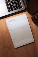 A wooden desk holds a silver laptop with black keys, partially open, next to a notepad labeled 'Focus du jour'. The notepad has spaces for listing priorities and a daily schedule divided into morning, afternoon, and evening slots. A small portion of a brown cylindrical object, possibly a candle or cup, is visible to the right.