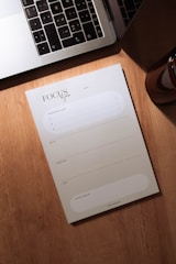 A wooden desk holds a silver laptop with black keys, partially open, next to a notepad labeled 'Focus du jour'. The notepad has spaces for listing priorities and a daily schedule divided into morning, afternoon, and evening slots. A small portion of a brown cylindrical object, possibly a candle or cup, is visible to the right.