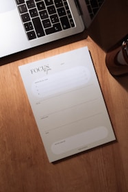 A wooden desk holds a silver laptop with black keys, partially open, next to a notepad labeled 'Focus du jour'. The notepad has spaces for listing priorities and a daily schedule divided into morning, afternoon, and evening slots. A small portion of a brown cylindrical object, possibly a candle or cup, is visible to the right.