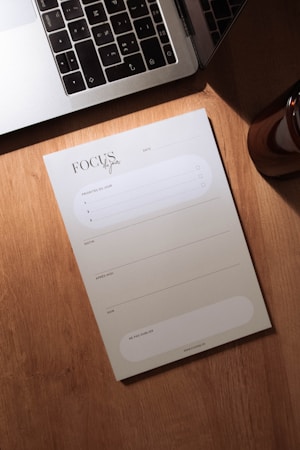 A wooden desk holds a silver laptop with black keys, partially open, next to a notepad labeled 'Focus du jour'. The notepad has spaces for listing priorities and a daily schedule divided into morning, afternoon, and evening slots. A small portion of a brown cylindrical object, possibly a candle or cup, is visible to the right.