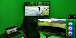 A gaming setup with a large curved monitor showing a scenic image of a river and forest. Several smaller monitors display nature scenes, and a laptop is open showing a different video. A comfortable gaming chair is positioned in front of the setup. The room is illuminated by green lighting, and the desktop tower has colorful RGB fans.