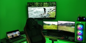 A gaming setup with a large curved monitor showing a scenic image of a river and forest. Several smaller monitors display nature scenes, and a laptop is open showing a different video. A comfortable gaming chair is positioned in front of the setup. The room is illuminated by green lighting, and the desktop tower has colorful RGB fans.