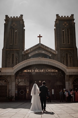 A couple, with the bride in a white gown and veil and the groom in a dark suit, stands side by side facing the entrance of Sacred Heart Church. The church features two tall towers and a central cross above the entrance. Several other people are gathered near the entrance.
