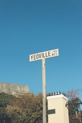 A street sign labeled 'Yeoville Rd/Weg' is mounted on a post against a clear blue sky. In the background, there is foliage and part of a white structure with a spiked fence. A cliff or rock formation is partially visible to the left.