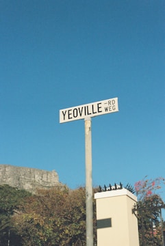 A street sign labeled 'Yeoville Rd/Weg' is mounted on a post against a clear blue sky. In the background, there is foliage and part of a white structure with a spiked fence. A cliff or rock formation is partially visible to the left.